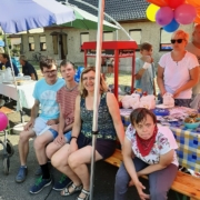 Vor dem Pop-Korn-Stand sitzen zwei Männer und eine Frau aus dem Heidehaus mit ihrer Betreuerin. Im Hintergrund sind die Pop-Korn-Maschine und Tütchen mit Pop-Korn zu sehen.