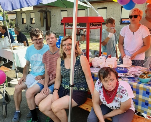 Vor dem Pop-Korn-Stand sitzen zwei Männer und eine Frau aus dem Heidehaus mit ihrer Betreuerin. Im Hintergrund sind die Pop-Korn-Maschine und Tütchen mit Pop-Korn zu sehen.