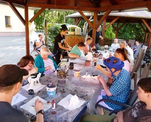 Die Bewohnerinnen und Bewohner des Sonnenhofes sitzen an langen Tischen und bemalen weiße Keramikfiguren.