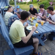 Die 7 Urlauber haben sich um einen blauen Holztisch versammelt und lassen sich das frisch Gegrillte und kühle Getränke schmecken.