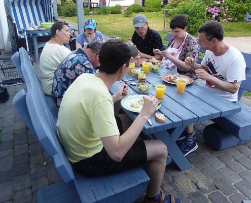 Die 7 Urlauber haben sich um einen blauen Holztisch versammelt und lassen sich das frisch Gegrillte und kühle Getränke schmecken.