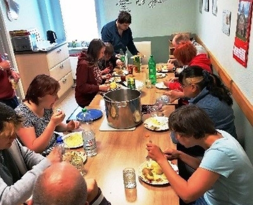 Alle Bewohner sitzen um einen großen Holztisch und essen. In der Mitte steht ein großer Edelstahltopf.