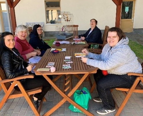 Mehrere Bewohnerinnen sitzen mit ihrer Betreuern unter einem Holz-Pavillon an einem Tisch. Unter anderem spielen sie Karten