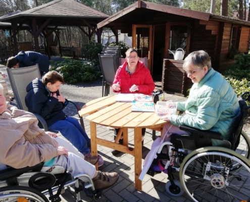 Vier Frauen, zwei davon im Rollstuhl, sitzen vor einem Gartenhäuschen an einem runden Holztisch in der Sonne. Sie basteln. Im Hintergrund limks von Ihnen bauen die beiden Männer den Jenga-Turm auf.