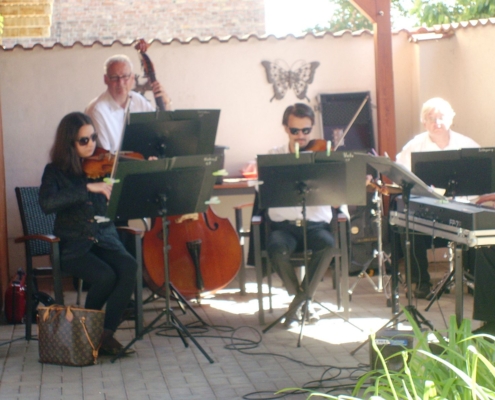 Eine Musikerin und drei Musiker sitzen unter einem Holzpavillon. Zwei von ihnen spielen Violine, einer Kontrabass und einer Trommel. Ganz rechts steht noch ein Keyboard. Vom Musiker sind nur die Hände zu sehen.