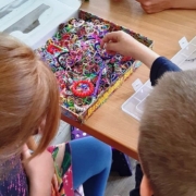 Zwei Mädchen und ein Junge sitzen am Tisch und basteln bunte Bänder. Zwischen ihnen steht ein Karton mit vielen Einzelteilen.