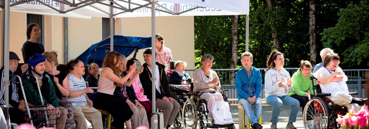 Teilhabe statt Ausgrenzung_Lebenshilfe_Barnim Viele Männer und Frauen sitzen auf Bänken oder in ihren Rollstühlen, teilweise unter einem Pavillon. Am Rand des Pavillondaches steht vorne und hinten: #TeilhabeStattAusgrenzung. Links und reicht ist Lebenshilfe Barnim zu lesen.