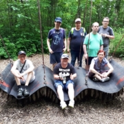 Auf einer breiten Schaukel, die aussieht wie ein fliegender Teppich und an acht Seilen befestigt ist, sitzen eine Frau und zwei Männer. Dahinter stehen eine weitere Frau und drei Männer. Die Schaukel befindet sich auf einem freien Platz im Wald.