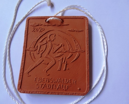 Die Stadtlaufmedaille ist rechteckig aus braunem Ton mit einer weißen Kordel. Auf der Platte sind ein stilisierter Läufer, das Jahr 2020 und die Beschriftung 14. Eberswalder Stadtlauf als Relief abgebildet.