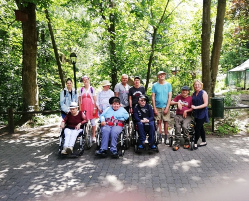 Die Gruppe hat sich am Rande eines Geheges, das teilweise mit Bäumen bewachsen ist, zum Gruppenfoto aufgestellt. Vorn stehen drei Rollstuhlfahrer und eine Person mit Rollator. Dahinter stehen die sieben anderen Teilnehmer.