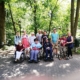 Die Gruppe hat sich am Rande eines Geheges, das teilweise mit Bäumen bewachsen ist, zum Gruppenfoto aufgestellt. Vorn stehen drei Rollstuhlfahrer und eine Person mit Rollator. Dahinter stehen die sieben anderen Teilnehmer.