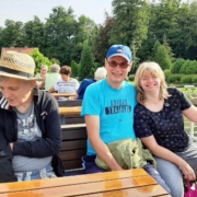 Auf dem Oberdeck der "Altwarp" sitzen drei der Ausflügler ,eine Frau und zwei Männer, auf einer dunkelbraunen Holzbank an einem Tisch. Der eine Mann hat seine linken Arm um die Freu gelegt. Das Paar lacht in die Kamera. Im Hintergrund ist das von Bäumen bestandene Ufer zu sehen.