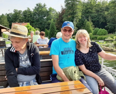 Auf dem Oberdeck der "Altwarp" sitzen drei der Ausflügler ,eine Frau und zwei Männer, auf einer dunkelbraunen Holzbank an einem Tisch. Der eine Mann hat seine linken Arm um die Freu gelegt. Das Paar lacht in die Kamera. Im Hintergrund ist das von Bäumen bestandene Ufer zu sehen.