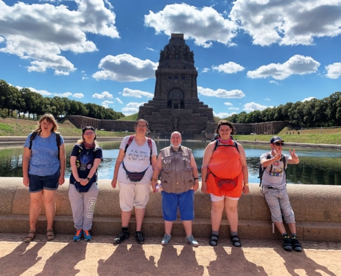 Fünf Frauen und ein Mann aus der Reisegruppe stehen am Rand des Wasserbeckens. Im Hintergrund ragt das Völkerschlachtdenkmal in den blauen Himmel mit kleinen weißen Wolken.