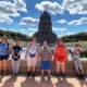 Fünf Frauen und ein Mann aus der Reisegruppe stehen am Rand des Wasserbeckens. Im Hintergrund ragt das Völkerschlachtdenkmal in den blauen Himmel mit kleinen weißen Wolken.