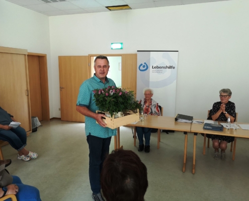 Manfred Groth steht mit einer Stiege mit Blumen in beiden Händen. Er wendet sich den Mitgliedern zu.