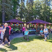 Am Rand der Wiese stehen zwei Holz-Pavillons. Diese sind mit bunten Girlanden und Luftballons geschmückt. Darunter und davor stehen und siezten die Bewohnerinnen und Bewohner. Sie unterhalten sich.