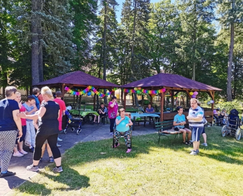 Am Rand der Wiese stehen zwei Holz-Pavillons. Diese sind mit bunten Girlanden und Luftballons geschmückt. Darunter und davor stehen und siezten die Bewohnerinnen und Bewohner. Sie unterhalten sich.
