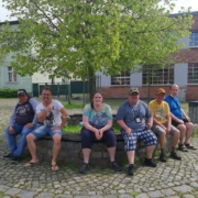 Fünf Männer und ein junge Frau aus der Wohngemeinschaft Breite Straße 21 sitzen auf der runden Einfassung eines Baumes auf dem Eberswalder Richterplatz. Die meisten tragen T-Shirts und kurze Hosen. Im Hintergrund ist noch ein Teil des Gebäudes der Wohngemeinschaft Mauerstraße 21 zusehen.