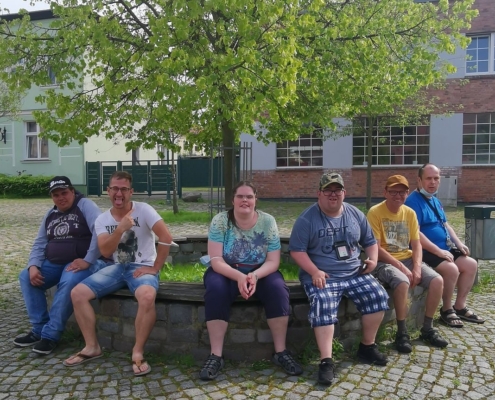 Fünf Männer und ein junge Frau aus der Wohngemeinschaft Breite Straße 21 sitzen auf der runden Einfassung eines Baumes auf dem Eberswalder Richterplatz. Die meisten tragen T-Shirts und kurze Hosen. Im Hintergrund ist noch ein Teil des Gebäudes der Wohngemeinschaft Mauerstraße 21 zusehen.