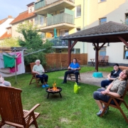 Vier Bewohnerinnen und ein Bewohner sitzen auf Stühlen im Kreis um eine Feuerschale im Hof der Jüdenstraße 7. Ein Stuhl im Vordergrund ist frei. Neben der Feuerschale steht eine hellgrüne Plastegießkanne. Auf einer Wäschespinne hängt Wäsche.