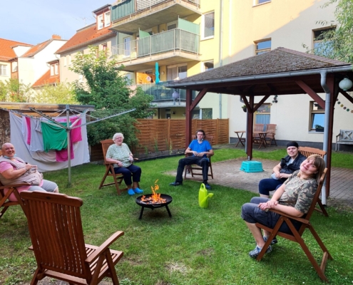 Vier Bewohnerinnen und ein Bewohner sitzen auf Stühlen im Kreis um eine Feuerschale im Hof der Jüdenstraße 7. Ein Stuhl im Vordergrund ist frei. Neben der Feuerschale steht eine hellgrüne Plastegießkanne. Auf einer Wäschespinne hängt Wäsche.