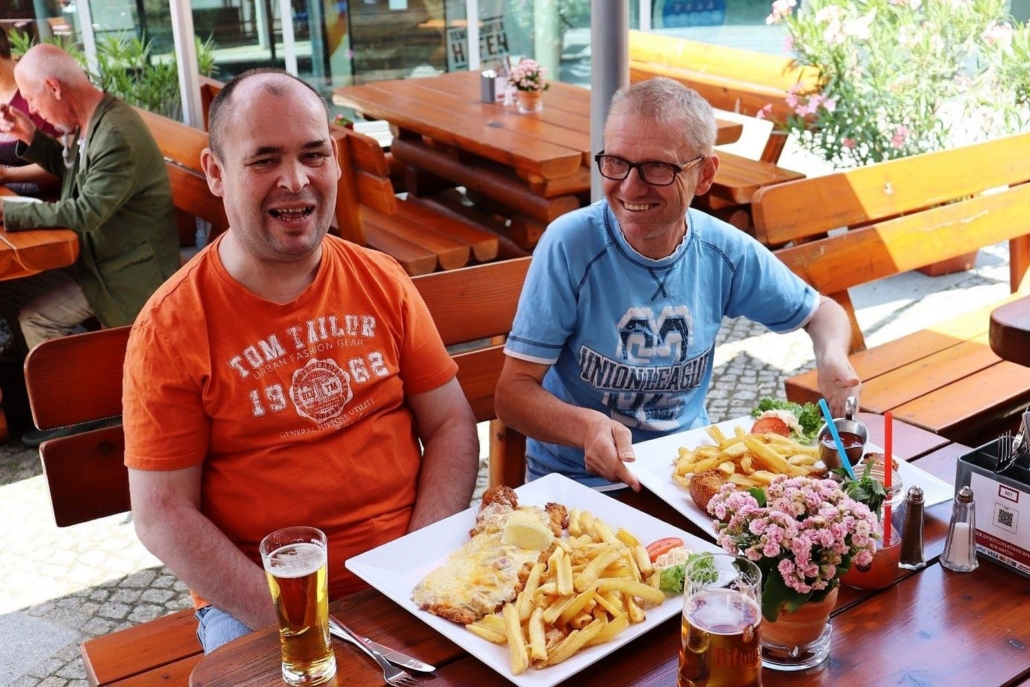 FuBs_Sommer 2021: Ob wir das alles schaffen? Zwei Männer sitzen im REDO XXL Restaurant. Vor sich haben sie jeder einen riesige Portion Pommes frites mit einem XXL-Schnitzel. Beide lachen.