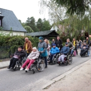 Auf der Straße des Dr.-Zinn-Weges läufen und rollen die Bewohnerinnen und Bewohner der Wohnstätte Sonnenhof, angeführt von sieben Rollifahrern mit ihren Begleitern der Deutschen Post. Alle sind warm angezogen. auf der linken Seite sind Einfamilienhäuser zusehen.