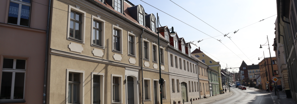 Ab der Breite Straße 54 ist die Straße in Richtung Ostende abgebildet. Links vorn befindet sich die das Haus 54 mit der Wohngemeinschaft. Es hat mit Dach- und Erdgeschoss drei Etagen. Im Dachgeschoss sind fünf Gauben. Die Fassade ist beige geputzt.