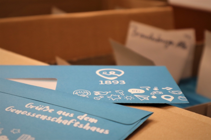 Die Fensterumschläge der Wohnungsgenossenschaft 1893. Auf dem Foto sind Vor- und Rückseite der Briefumschläge der Wohnungsgenossenschaft 1893 abgebildet. Auf der Vorderseite sind das Logo und weitere Symbole aufgedruckt. Die Rückseite hat in weißer Schrift den Aufdruck "Grüße aus dem Genossenschaftshaus".