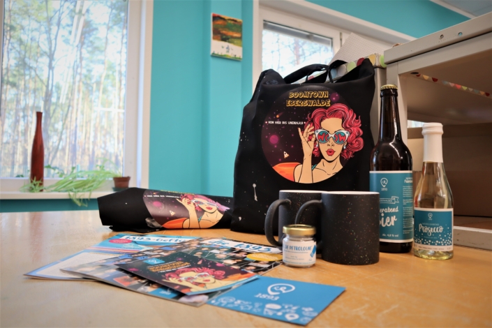 Ein Willkommensgruß für die neuen Mieter:Innen im Projekt Brand.Vier der 1893. Zu dem Willkommensgruß gehören unter anderem ein dunkler Stoffbeutel mit dem farbigen Logo Boomtown Eberswalde, eine kleine Flasche "Feierabendbier", eine kleine Flasche Prosecco, beide Etiketten in den Farben der 1893, zwei dunkle Tassentöpfe und verschiedene Info-Materialien.