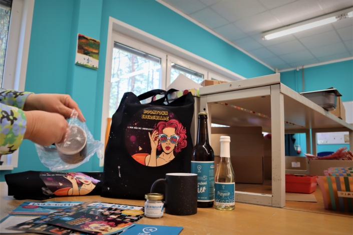 Der Willkommensgruß wird eingepackt. Eine Beschäftigte steckt einen dunklen Tassentopf in eine Plastiktüte. Auf dem Tisch stehen und liegen die anderen Bestandteile des Willkommensgrußes: ein dunkler Stoffbeutel mit dem farbigen Logo Boomtown Eberswalde, eine kleine Flasche "Feierabendbier", eine kleine Flasche Prosecco, beide Etiketten in den Farben der 1893, ein weiterer dunkler Tassentopf und verschiedene Info-Materialien.
