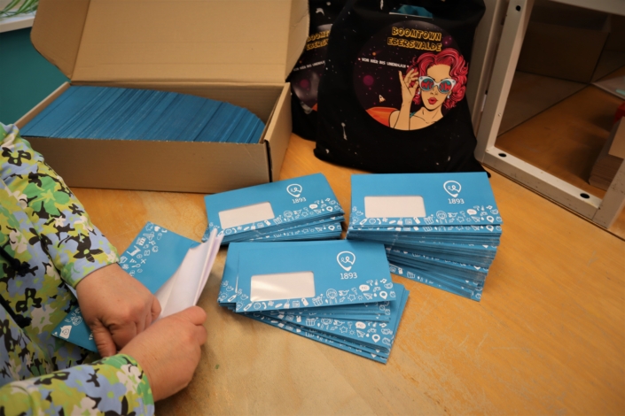 Die Briefe müssen in den Umschlag Eine Beschäftigte steckt die Briefe in blaue Fensterumschläge der Wohnungsgenossenschaft 1893. Am Rand steht ein fast voller Karton mit Briefumschlägen.