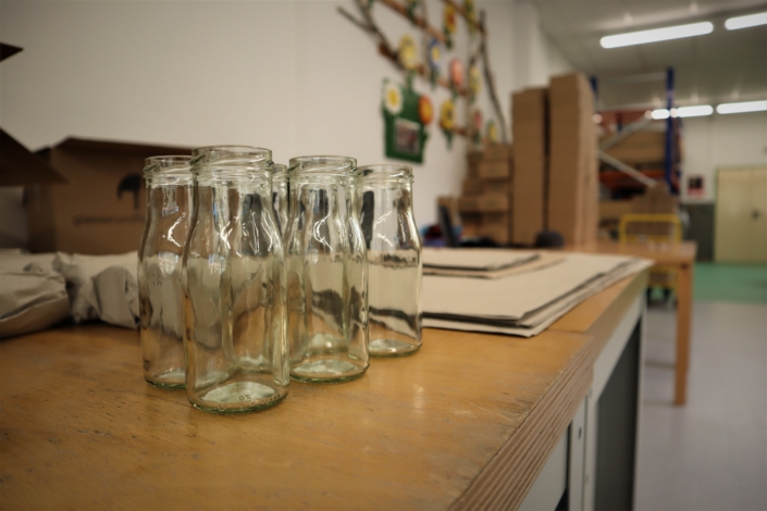 Die Glasflaschen stehen zum Verpacken bereit. Auf einem Arbeitstisch stehen fünf kleine Glasflaschen. Dahinter liegen bereits verpackte und ungefaltete Kartons.