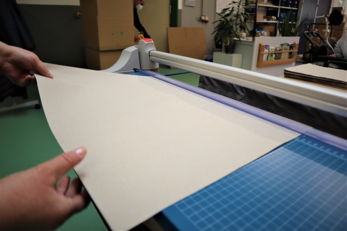 Zuschneiden des Packpapiers An die Schneidekante einer blauen Papierschneidemaschine wird ein Bogen Packpapier angelegt. Im Hintergrund sind an der Wand Regale zu sehen.