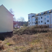 Das Baugrundstück ist mit trockenem Gras und trockenen, alten Stauden bewachsen. Links schließt sich der Giebel eines älteren Hauses an. Auf der rechten Seite sind relativ neugebaute Wohnhäuser sichtbar.