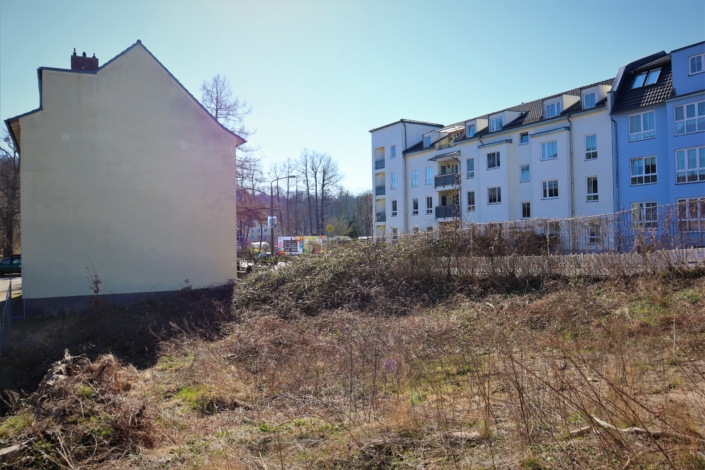 Baubeginn Raumerstraße – hier entsteht die neue Wohnstätte Das Baugrundstück ist mit trockenem Gras und trockenen, alten Stauden bewachsen. Links schließt sich der Giebel eines älteren Hauses an. Auf der rechten Seite sind relativ neugebaute Wohnhäuser sichtbar.