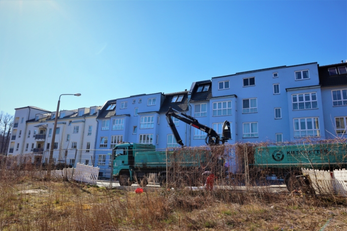 Baubeginn Raumerstraße – zum Entladen bereit Der große, grüne LKW mit integriertem Bagger und Anhänger der Firma Kiesewetter steht vor dem unbebauten Grundstück in der Raumerstraße. An der Längsseite des Anhängers ist das Logo der Firma zu sehen. Den Hintergrund bilden beige und blaue neugebaute Wohnhäuser.