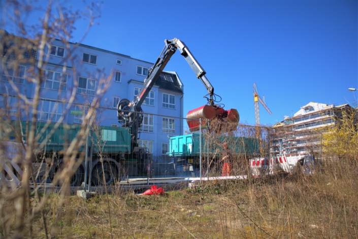 Baubeginn Raumerstraße – der LKW wird entladen Der Anhänger eines großen, grünen LKW wird mit einem Bagger entladen. Der Bagger ist am Heck des LKW montiert. Ein Bauarbeiter bedient den Bagger, ein zweiter weist ihn ein.