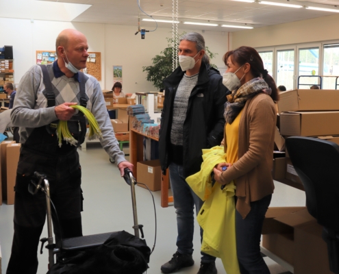 Frau Wiese und Herr Gloger sprechen in der Abteilung Montage mit dem Vorsitzenden des Werkstattrates. Dieser hält konfektionierte gelbe Kabel in seiner rechten Hand. Er stützt sich auf einen Rollator. Im Hintergrund sind andere Beschäftigte an ihren Arbeitsplätzen zu sehen.