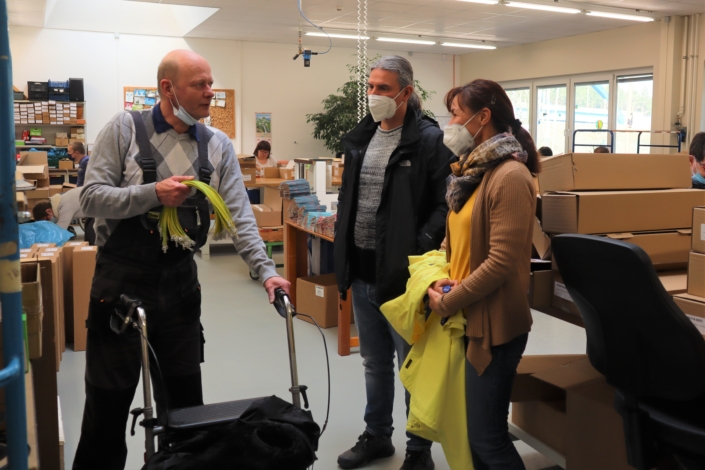 Frau Wiese und Herr Gloger sprechen in der Abteilung Montage mit dem Vorsitzenden des Werkstattrates. Dieser hält konfektionierte gelbe Kabel in seiner rechten Hand. Er stützt sich auf einen Rollator. Im Hintergrund sind andere Beschäftigte an ihren Arbeitsplätzen zu sehen.