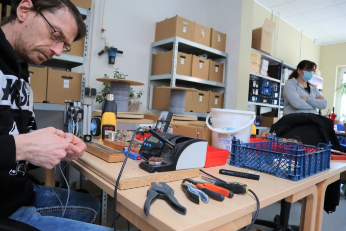 Auf einem Arbeitstisch aus hellem Holz stehen Messgeräte und ein Lötkolben. Am Rand liegen verschiedene Werkzeuge wie Zangen und Schraubenzieher. An der Wand stehen Regale mit Kisten. Ganz rechts ist die Gruppenleiterin Frau Urbick.