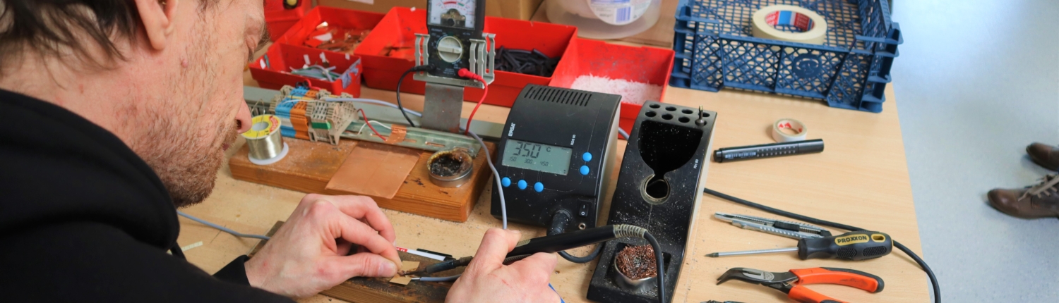 Ein Mitarbeiter lötet ein Kabel. Auf dem Tisch stehen verschiedene Messgeräte. Am Rand liegen Zangen und andere Werkzeuge.