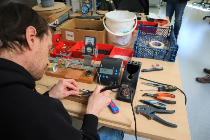Ein Mitarbeiter lötet ein Kabel. Auf dem Tisch stehen verschiedene Messgeräte. Am Rand liegen Zangen und andere Werkzeuge.