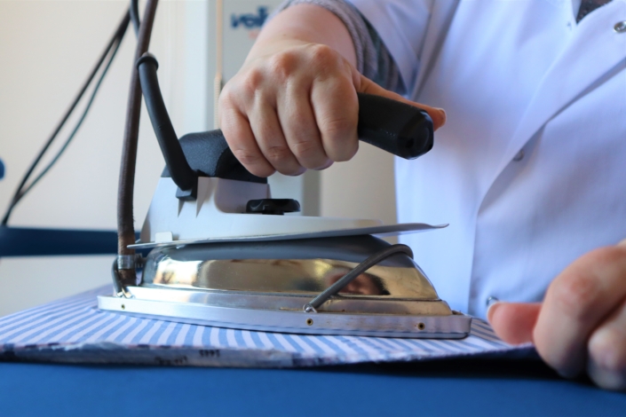 Im Mittelpunkt des Bildes steht eine Bügeleisen auf einem blau.-weiß gestreiften Tuch. Von der Büglerin sind nur die Hände und der Oberkörper zu sehen,