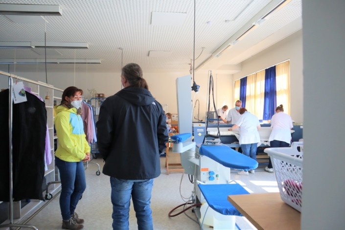 Ein Blick in den Bügelbereich der Wäscherei. Rechts stehen Bügelstationen und eine Mangel, an der drei Beschäftigte arbeiten. Im Vordergrund stehen Frau Wiese und Herr Gloger.