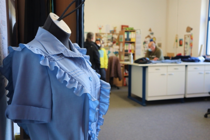 Auf einer Schneiderpuppe hängt eine blaue, kurzärmelige Bluse mit Rüschen am Vorderteil. Im Hintergrund ist ein Schreibtisch und eine Frau und zwei Männer zu sehen.