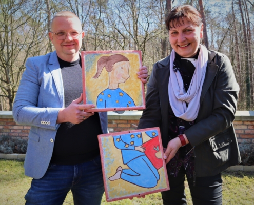 Herr Lutter (links im Bild) und Frau Wucherpfennig (rechts) halten lächelnd zwischen sich jeder eine quadratische Fließe mit dem Oberkörper bzw. den Unterkörper der "Europa" in den Händen. Die „Europa“ ist hier ein Mädchen mit brünettem Pferdeschwanz und blauem Jogginganzug mit goldenen Sternen.