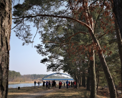 Das Bild wurde von der anderen Seite, aus größerer Entfernung aufgenommen. Die Sportlerinnen erwärmen sich mit verschiedenen Übungen,. Einige recken die Arme in die Luft. Im Hintergrund ist die blau-gelbe Kanalbrücke zu erkennbar.