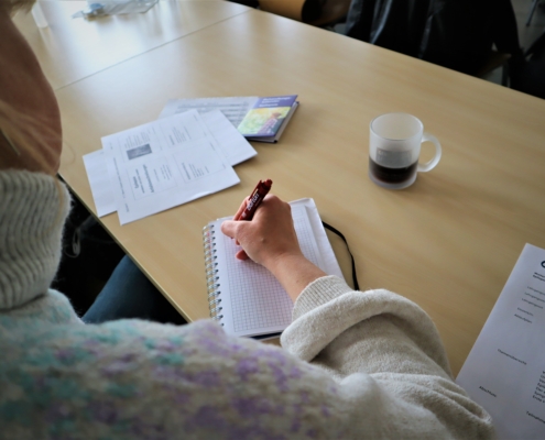 Eine Teilnehmerin macht sich in einem kleinen Ringbuch Notizen. Auf dem Tisch liegen verschiedene Unterlagen. Auch ein Tasse Kaffee steht dabei.